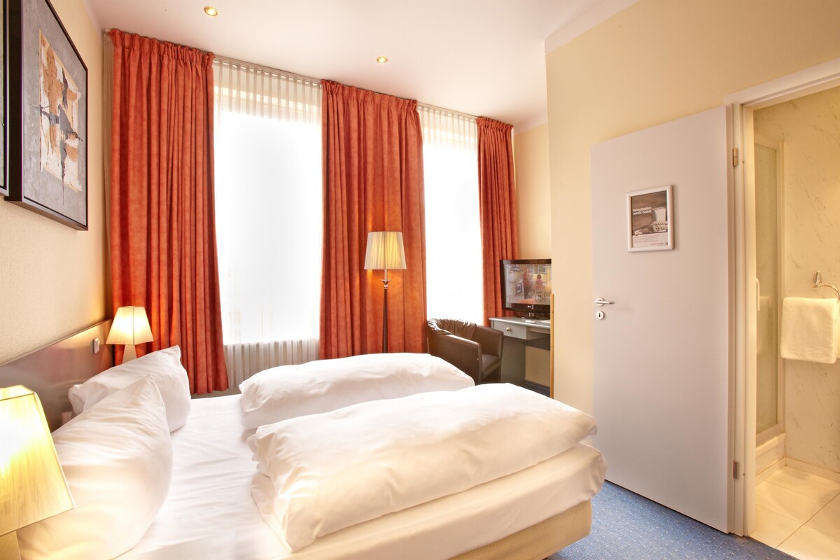 Ein Hotelzimmer mit zwei Betten, einem Schreibtisch, einem Stuhl, einer Lampe und einer Tür zu einem Badezimmer.