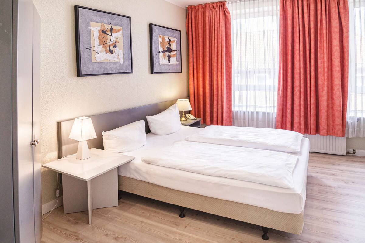 Ein Hotelzimmer mit Doppelbett, Nachttisch mit Lampe, zwei gerahmte abstrakte Gemälde an der Wand und einem Fenster mit roten Vorhängen.