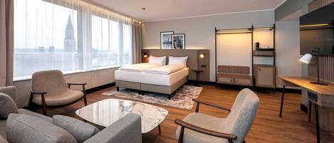 A modern hotel room with a large bed, a sofa, a coffee table, and a desk.