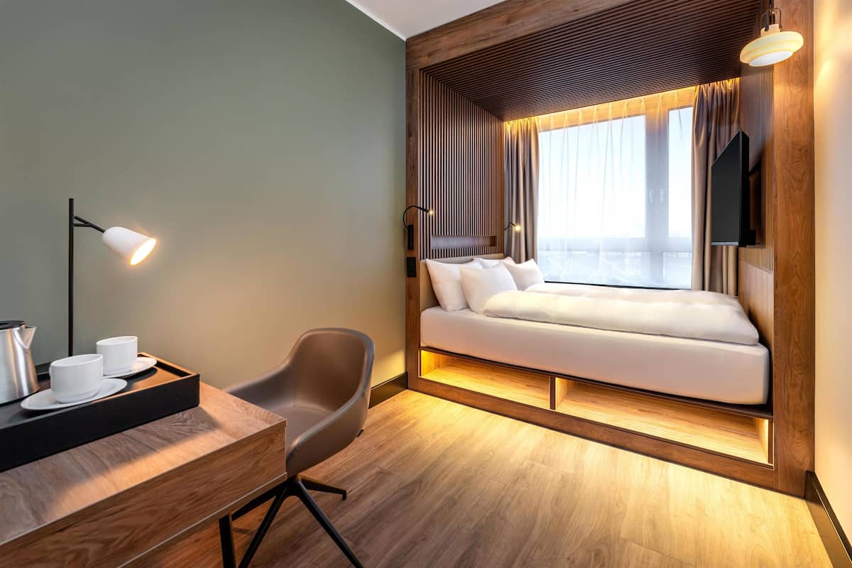 A modern hotel room with a wooden accent wall, a bed with white bedding, a desk with a lamp, and a TV mounted on the wall.