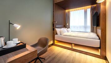 Una habitación de hotel moderna con un revestimiento de madera, una cama con ropa blanca, un escritorio con lámpara y un televisor montado en la pared.
