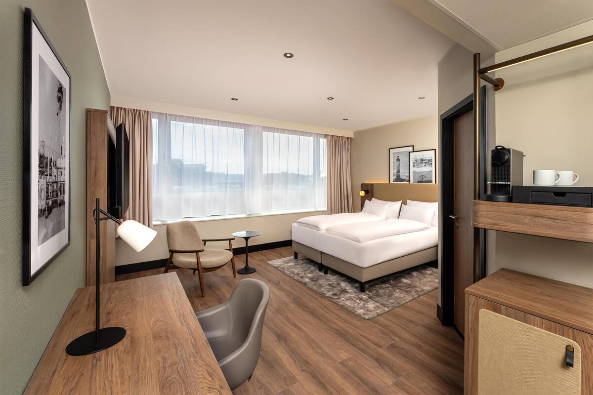 A hotel room with a bed, desk, chair, and a large window.