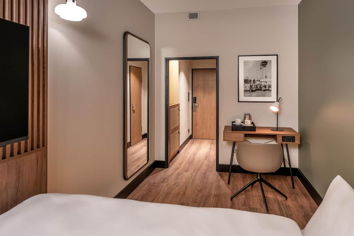 A hotel room with a bed, a desk with a chair, a mirror, a framed picture on the wall, and a wooden floor.
