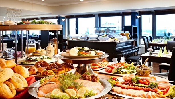 A buffet with various food items, a piano, and large windows with a view. - Hotel Astor Kiel by Campanile (Kiel)