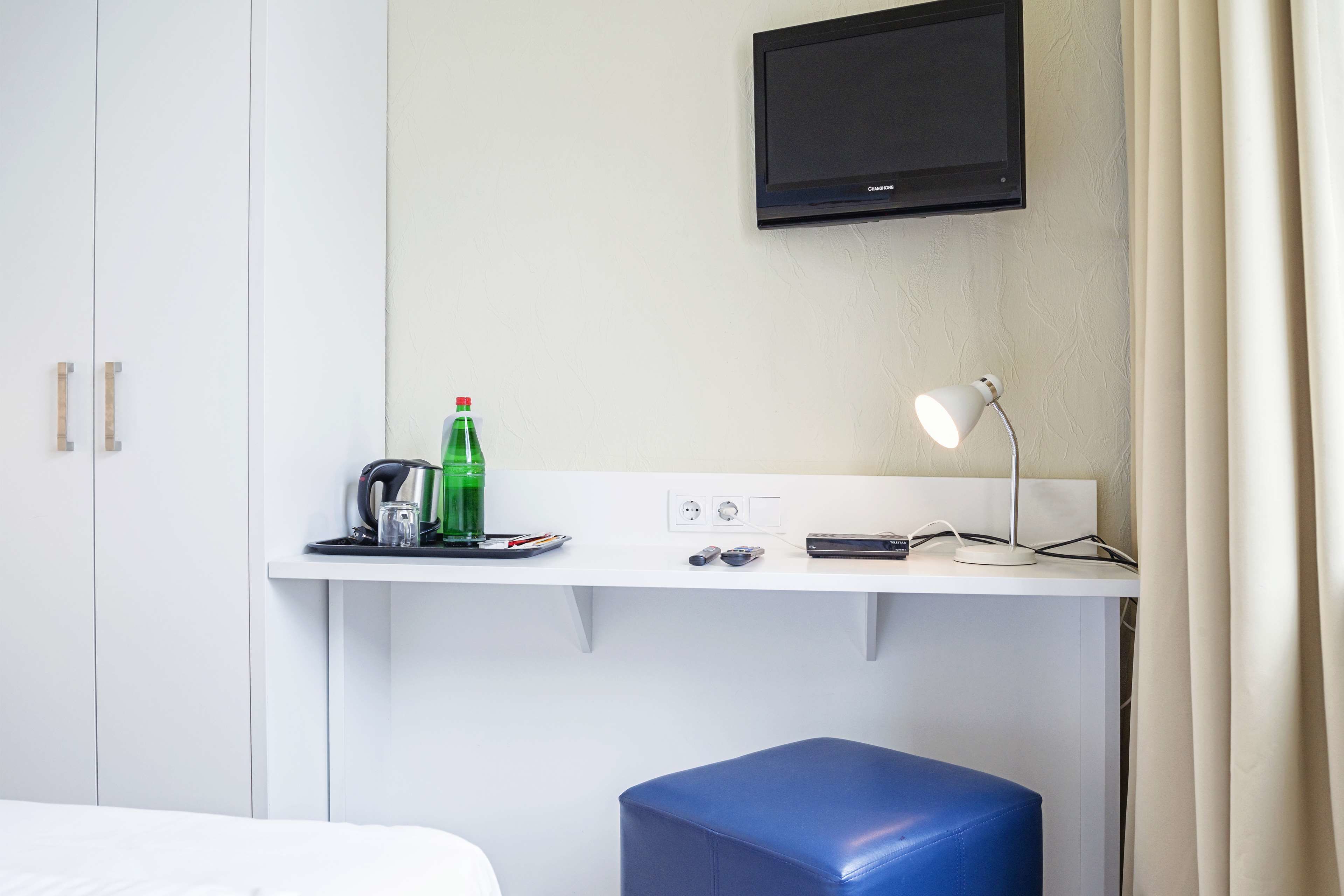 A hotel room with a white desk, a television mounted above it, a blue stool, and a bed with white bedding.