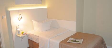 A neatly made bed with white linens, a bedside table with a bottle and a fruit, and a magazine on the bed.