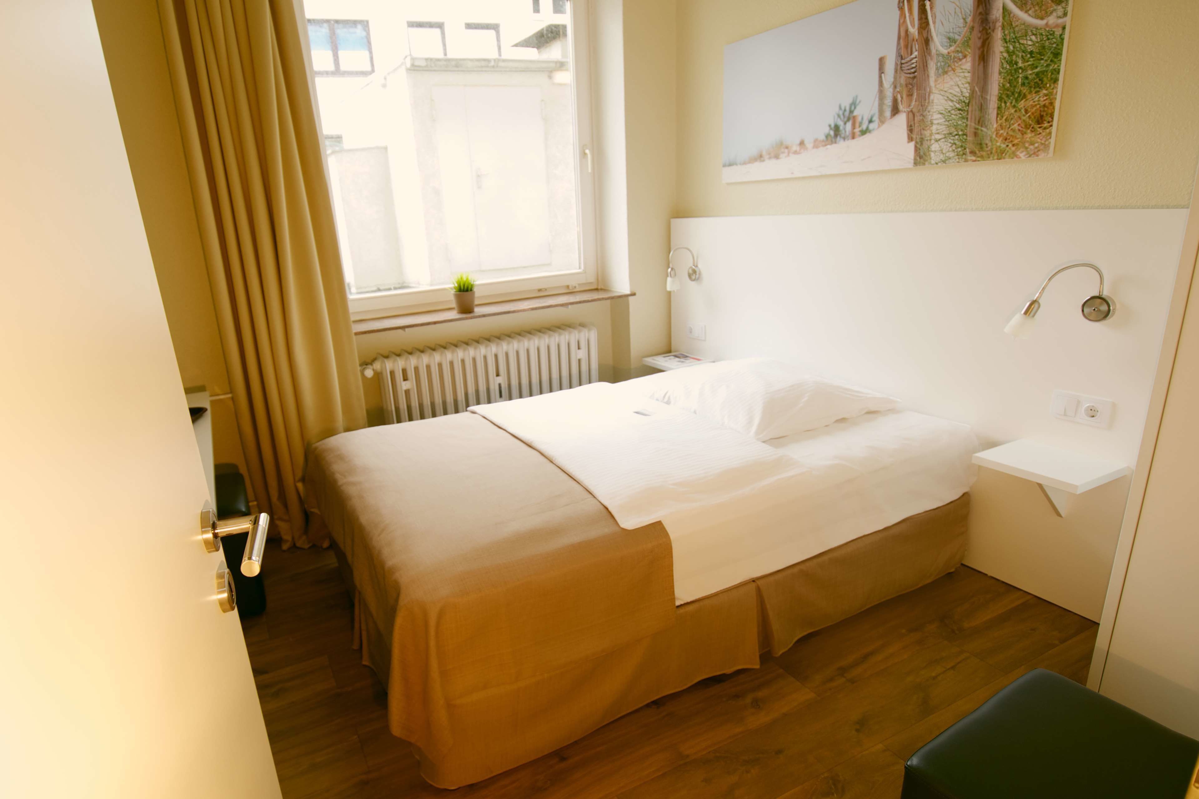 A hotel room with a bed, a window with curtains, a radiator, a bedside table, and a small green plant.