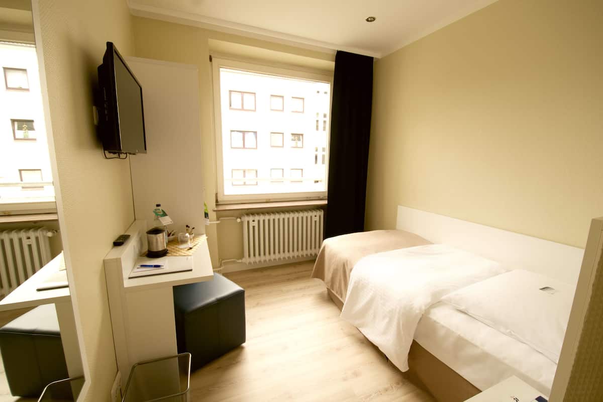 Une chambre d’hôtel comprenant un lit, un bureau avec un ordinateur, une chaise, une fenêtre avec des rideaux et un radiateur.