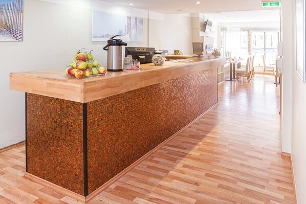 Zona de recepción de hotel con un mostrador de madera, una máquina de café y una exhibición de frutas.