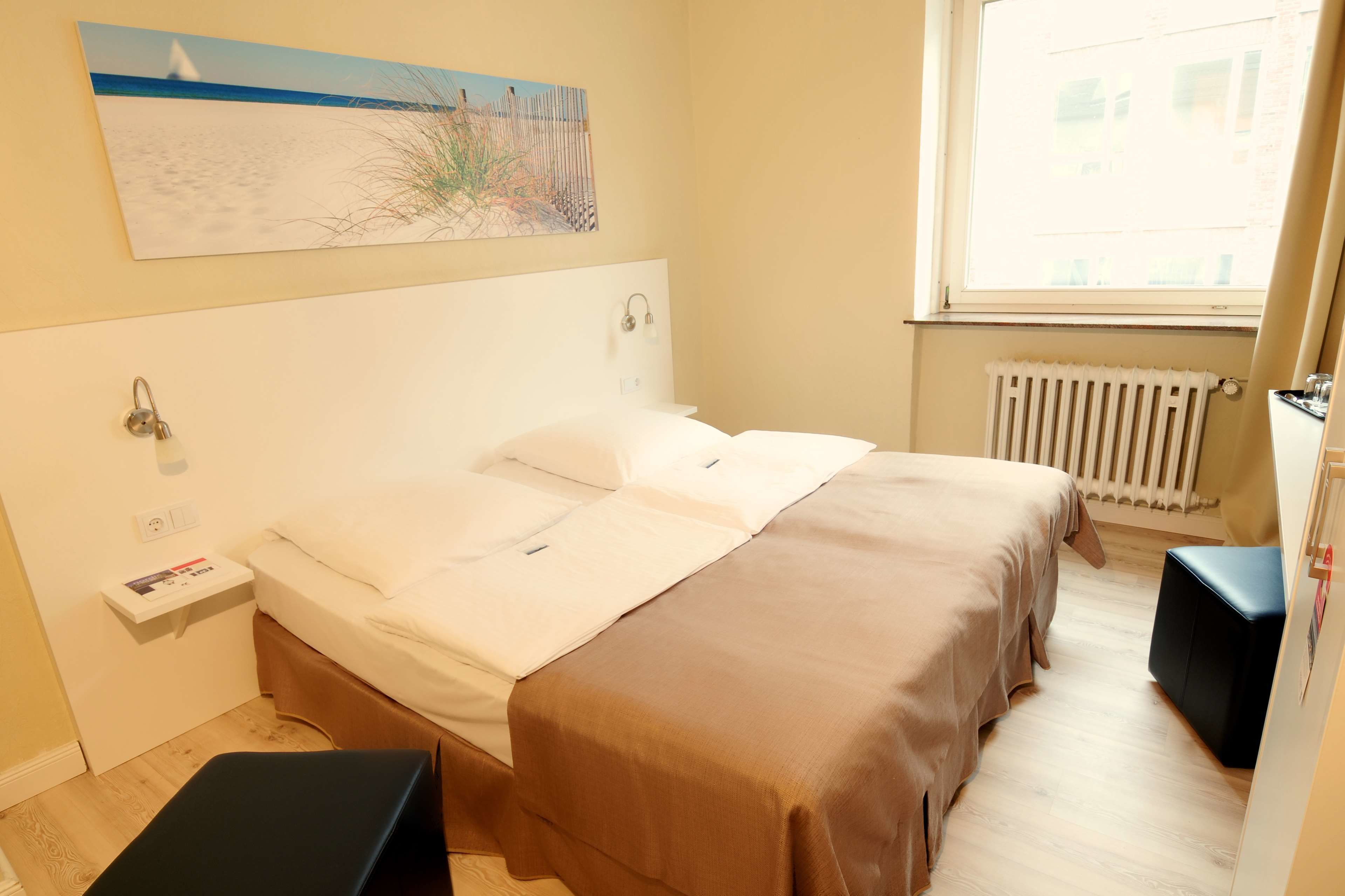 A double bed with a headboard, a bedside table with a lamp, a radiator, and a window with curtains.