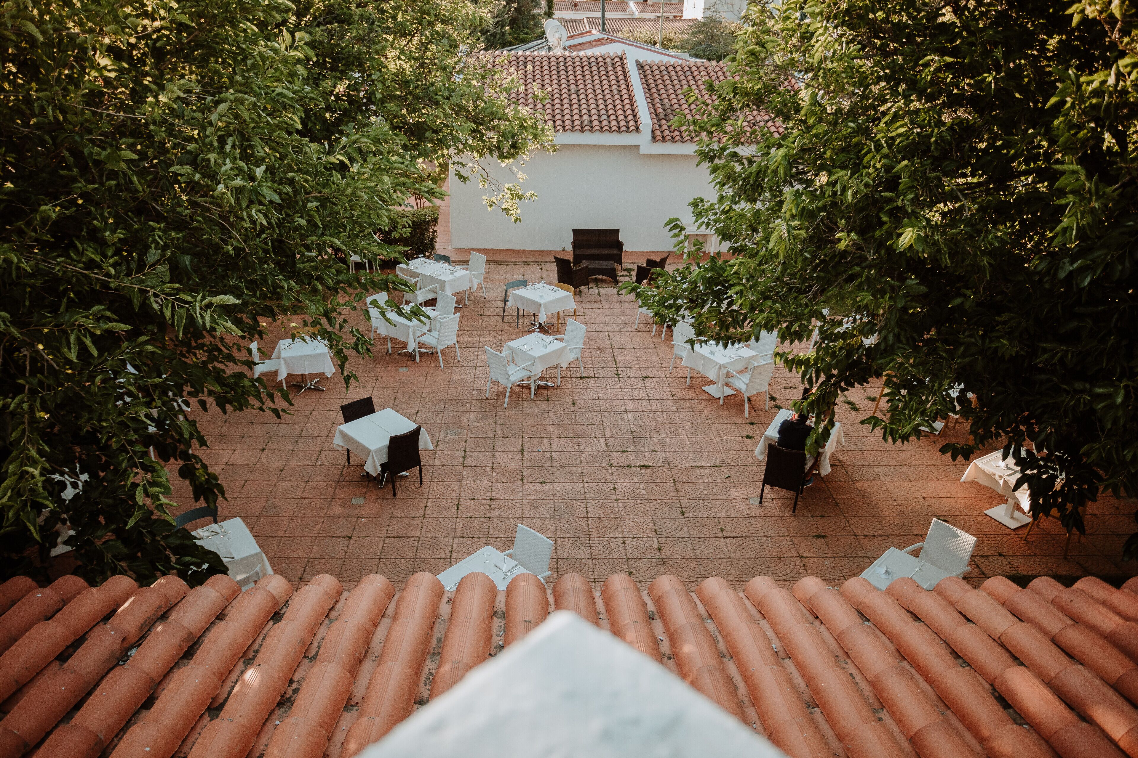 Un patio exterior con mesas y sillas blancas, rodeado de árboles.