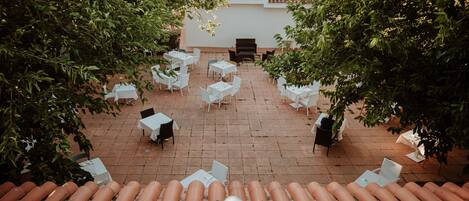 Un patio exterior con mesas y sillas blancas, rodeado de árboles.