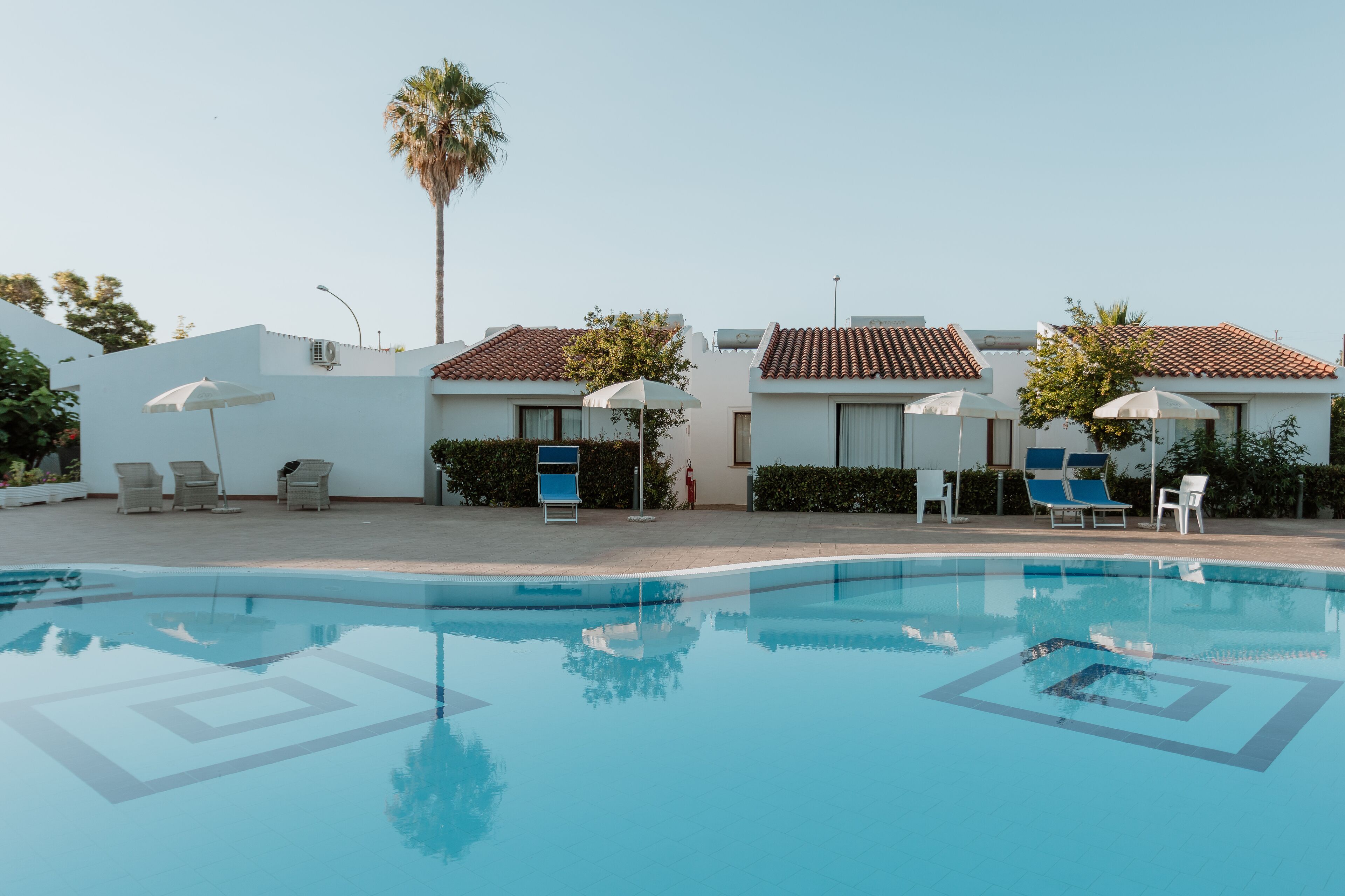 A swimming pool with lounge chairs and umbrellas, surrounded by buildings with tiled roofs and white walls.