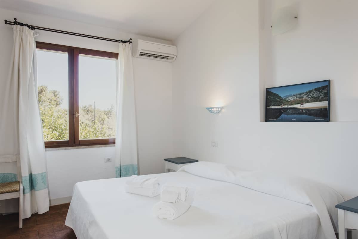 A hotel room with a bed, a nightstand, a window with curtains, an air conditioning unit, and a wall-mounted light fixture.