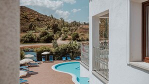 Un área de piscina con sillones y sombrillas, rodeada por un edificio blanco con balcón.