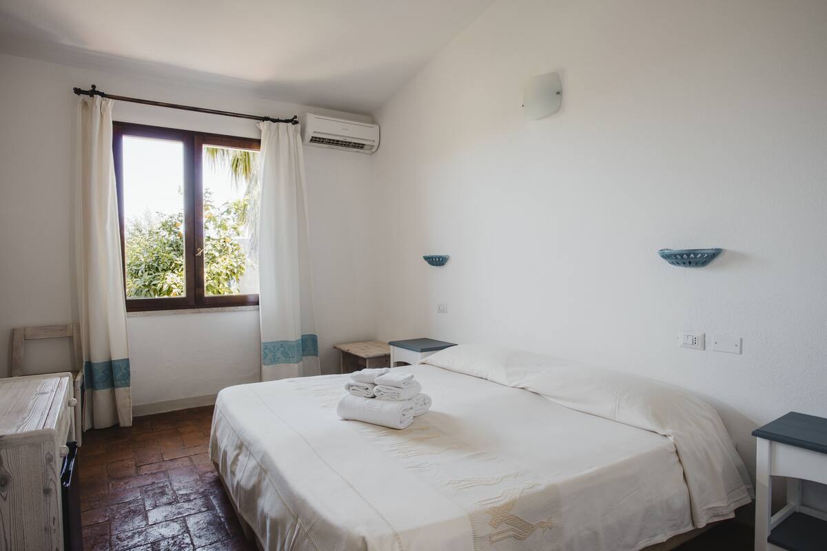 A bedroom with a bed, a window with curtains, a bedside table, and an air conditioning unit.