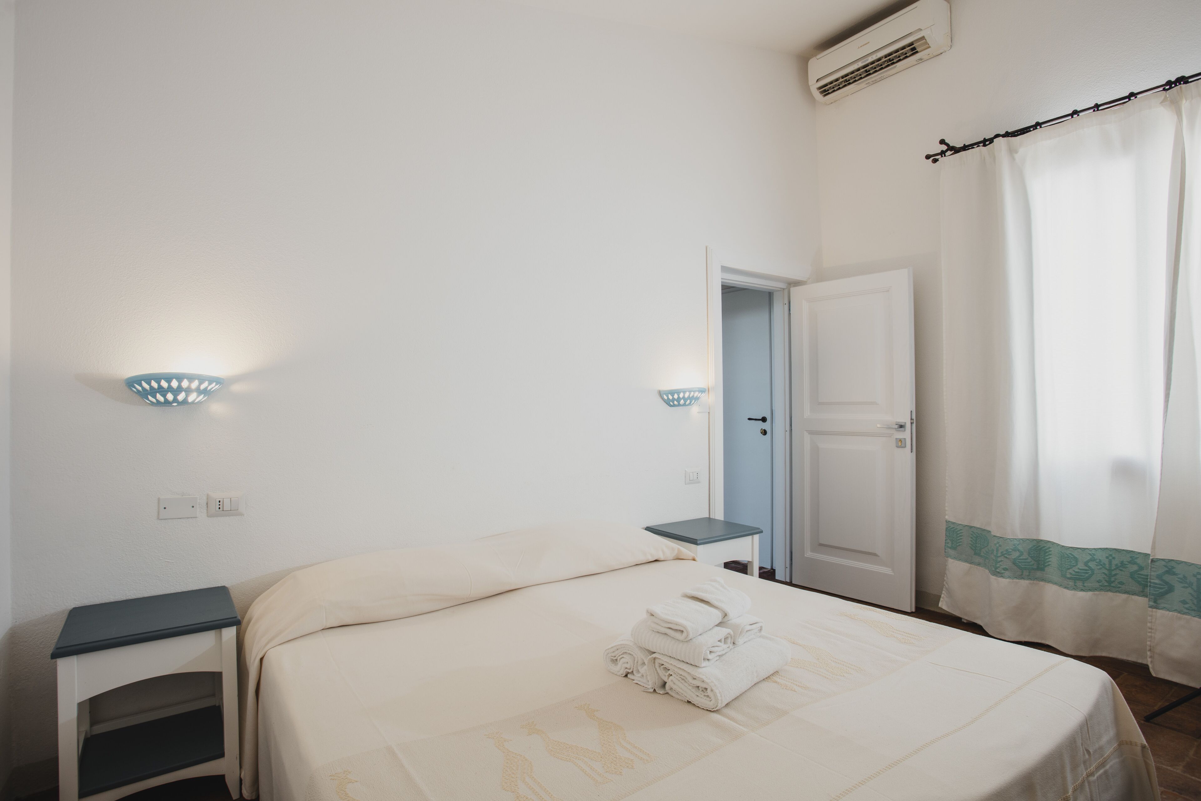 A hotel room with a bed, a nightstand, a wall-mounted light, a door, and a curtain.