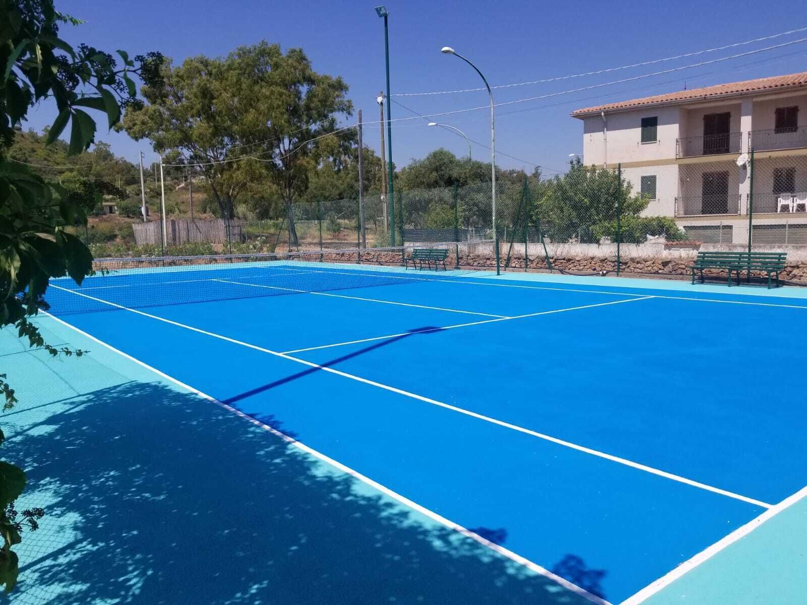 Una cancha de tenis con red, rodeada por una valla, árboles y un edificio con balcones.