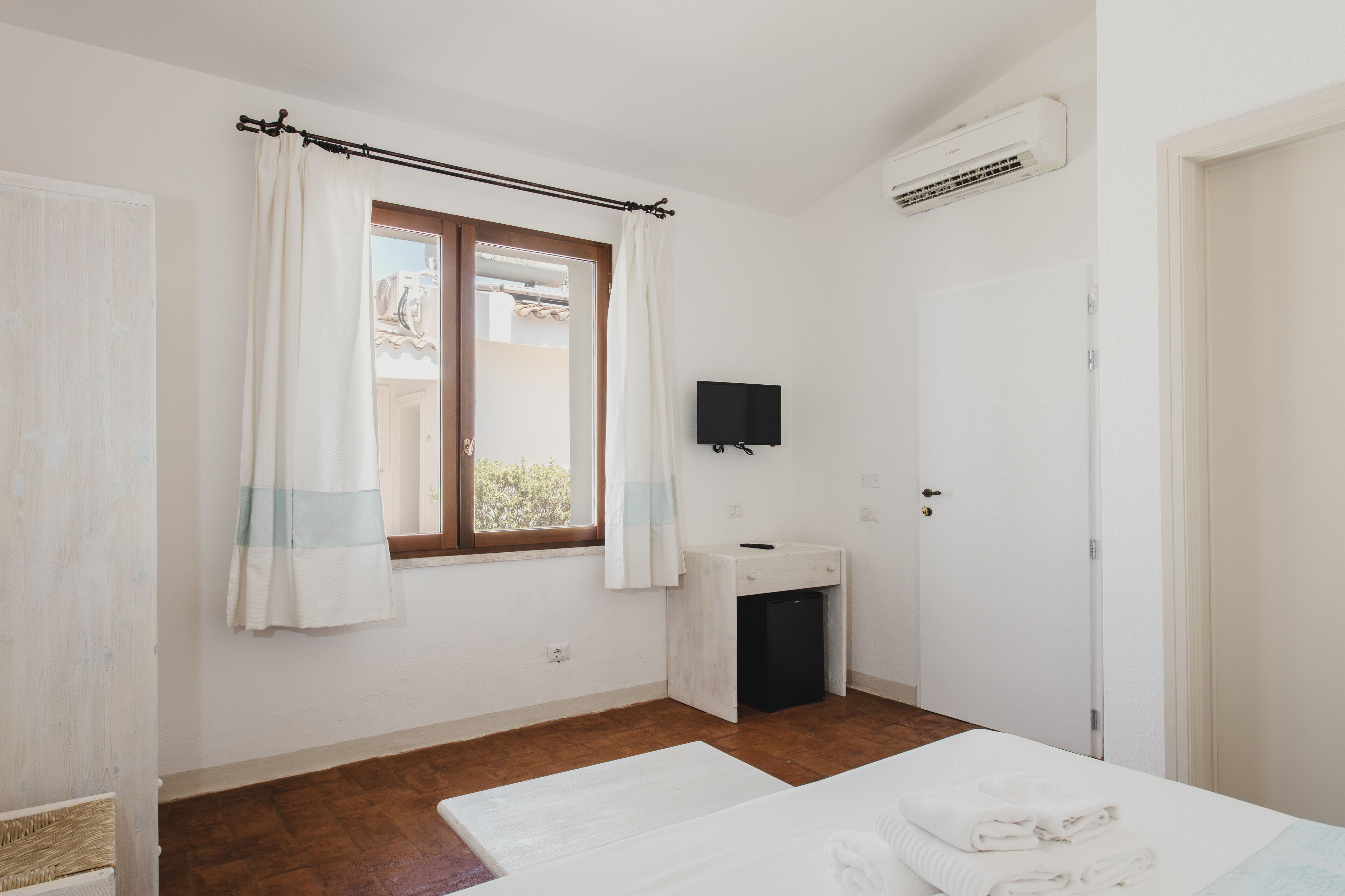 A bedroom with a bed, a small bedside table, a window with curtains, a wall-mounted TV, and an air conditioning unit.