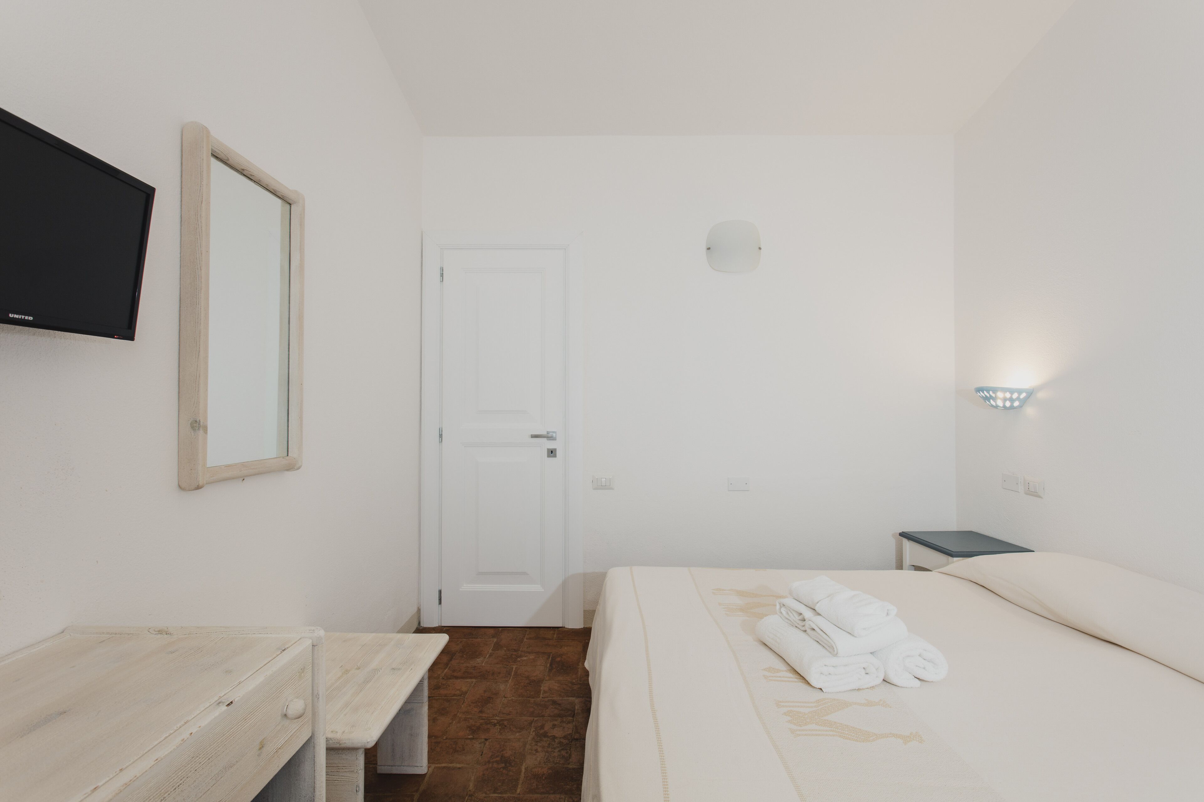 Une chambre d’hôtel avec un lit, un téléviseur fixé au mur, une porte et une petite table de chevet avec une lampe.
