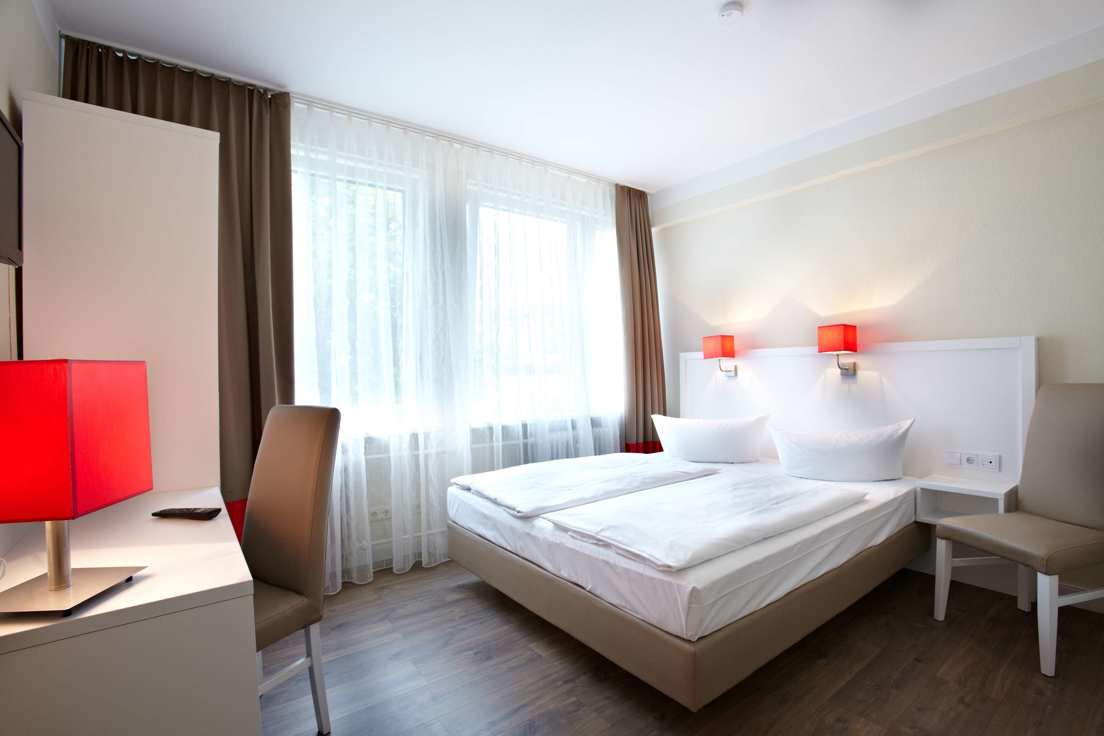 A hotel room with a bed, a desk with a lamp, a chair, and a nightstand with a lamp.