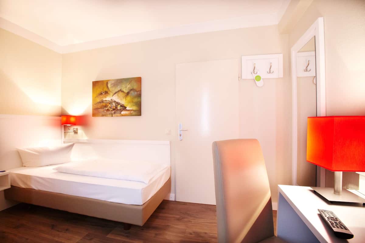 Une chambre d’hôtel avec un lit, un bureau doté d’une lampe, une chaise, un tableau accroché au mur et une porte.