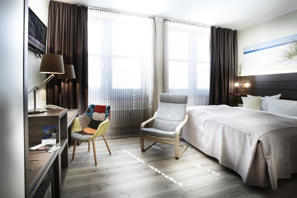 Une chambre d’hôtel avec un lit, un bureau, une chaise et une grande fenêtre avec des rideaux.