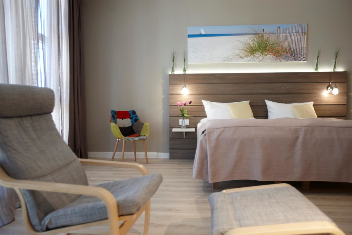 Une chambre d’hôtel moderne dotée d’un grand lit, d’un coin salon avec un fauteuil, une petite table et une œuvre d’art fixée au mur représentant une scène de plage.