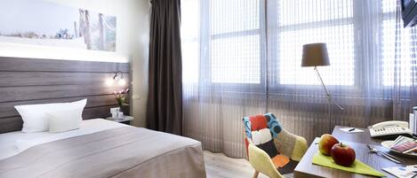 A hotel room with a bed, a desk with a lamp, a chair, and a window with curtains.