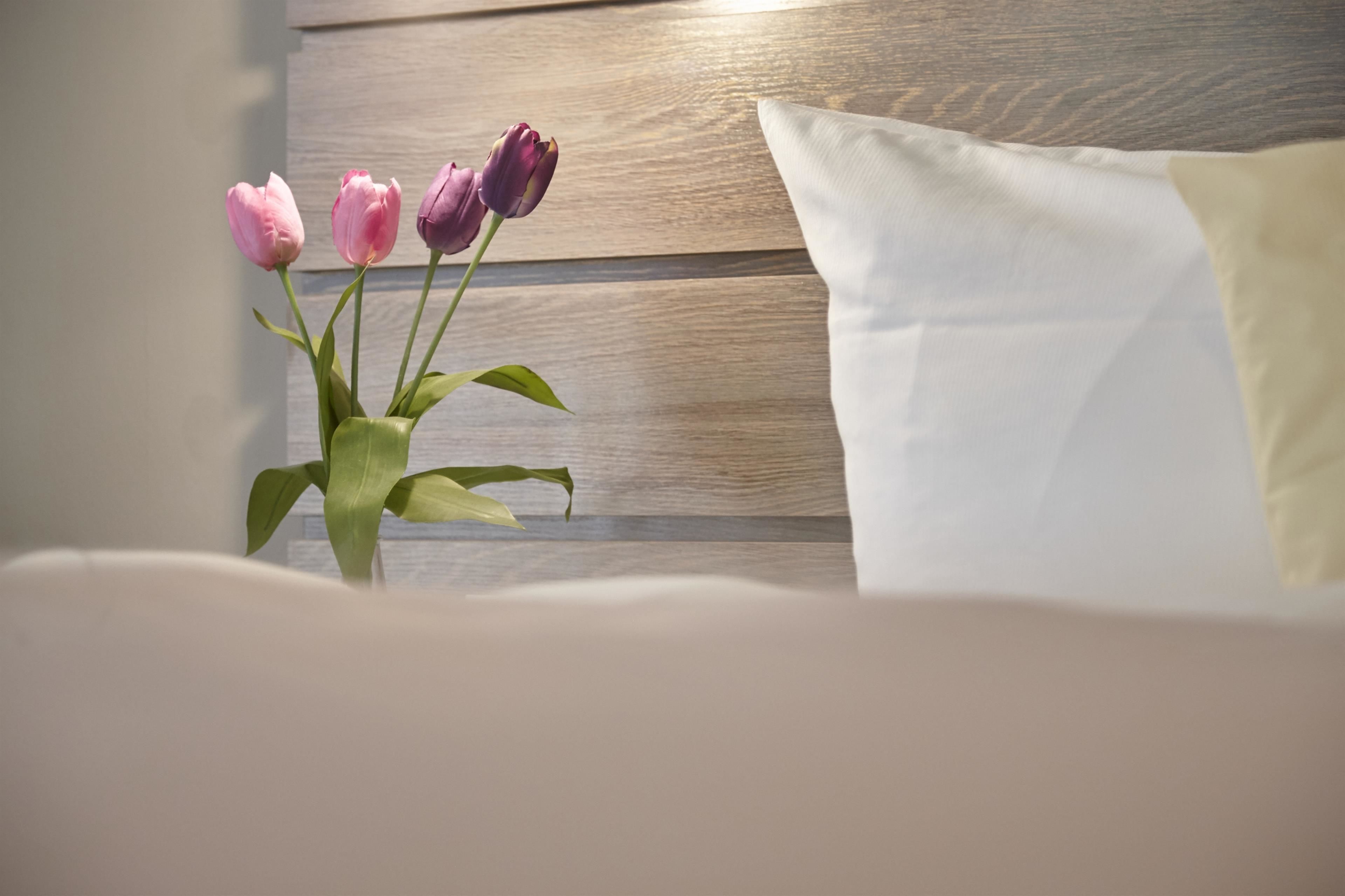 A bed with a white pillow and a vase with pink and purple flowers.