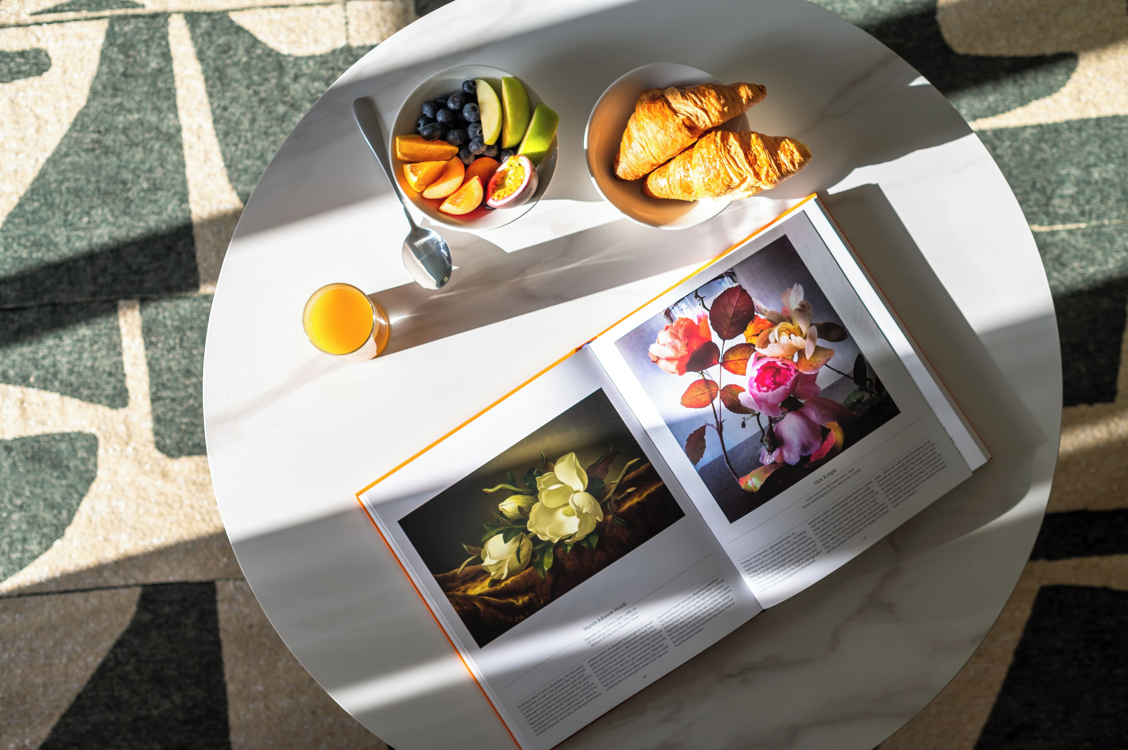 Una mesa redonda con mantel blanco, un cuenco de fruta, un croissant, un vaso de jugo de naranja y una revista abierta con motivos florales.