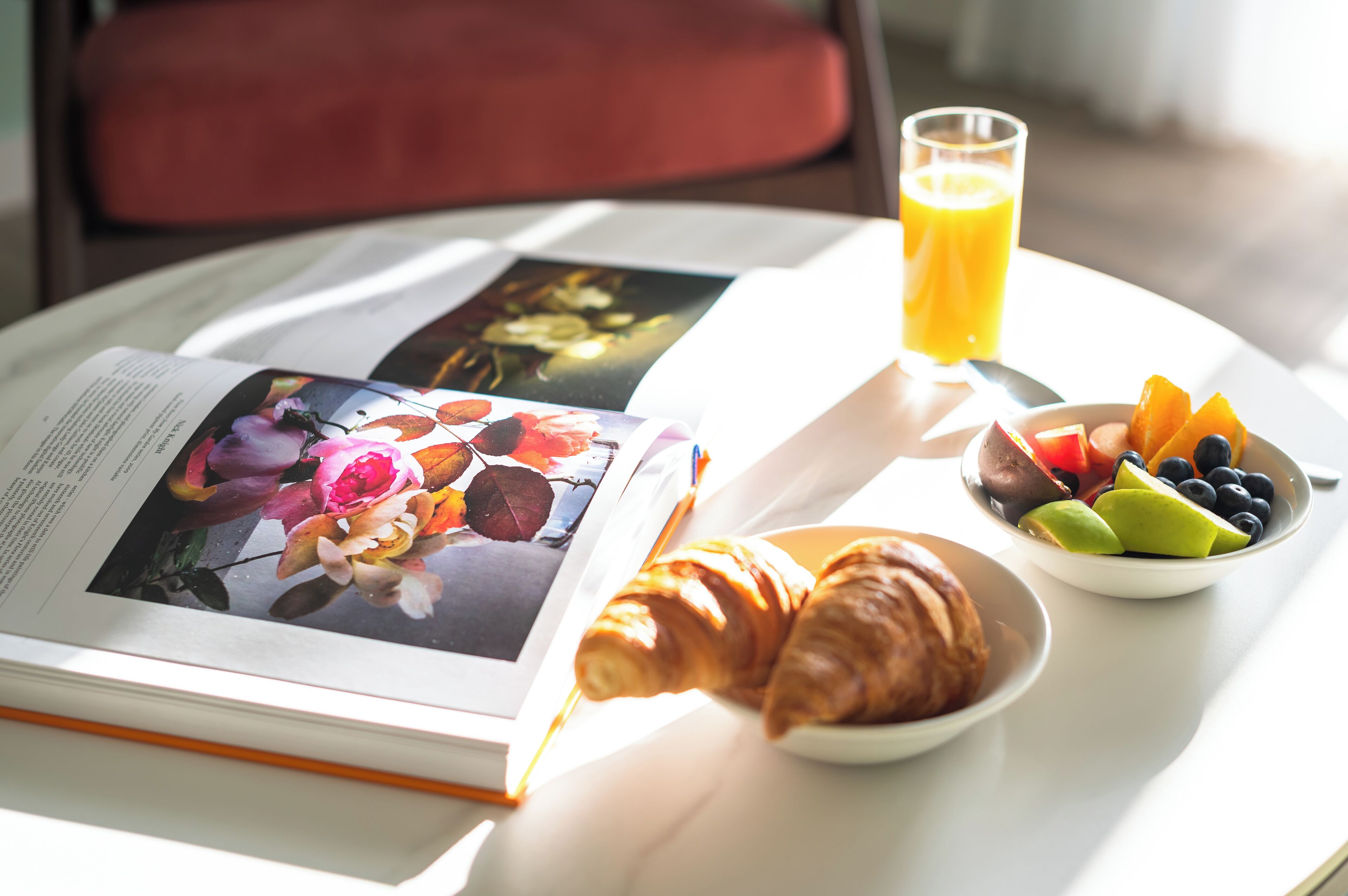 A breakfast setting with a book, croissants, a fruit bowl, and a glass of orange juice on a table.