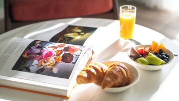 Un desayuno con un libro, croissants, un bol de fruta y un vaso de zumo de naranja sobre una mesa.