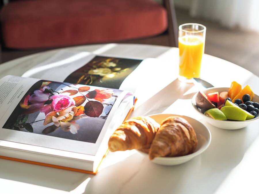 Un desayuno con un libro, croissants, un bol de fruta y un vaso de zumo de naranja sobre una mesa.