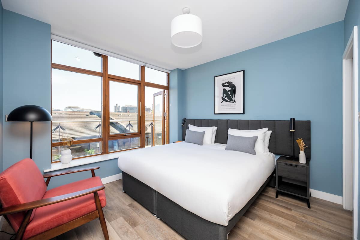 Een moderne slaapkamer met een groot bed, een rode fauteuil, een nachtkastje en een raam met uitzicht op de stad.