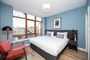 A modern bedroom with a large bed, a red chair, a nightstand, and a window with a city view. - Staycity Aparthotels, Dublin, Saint Augustine Street (Dublin)