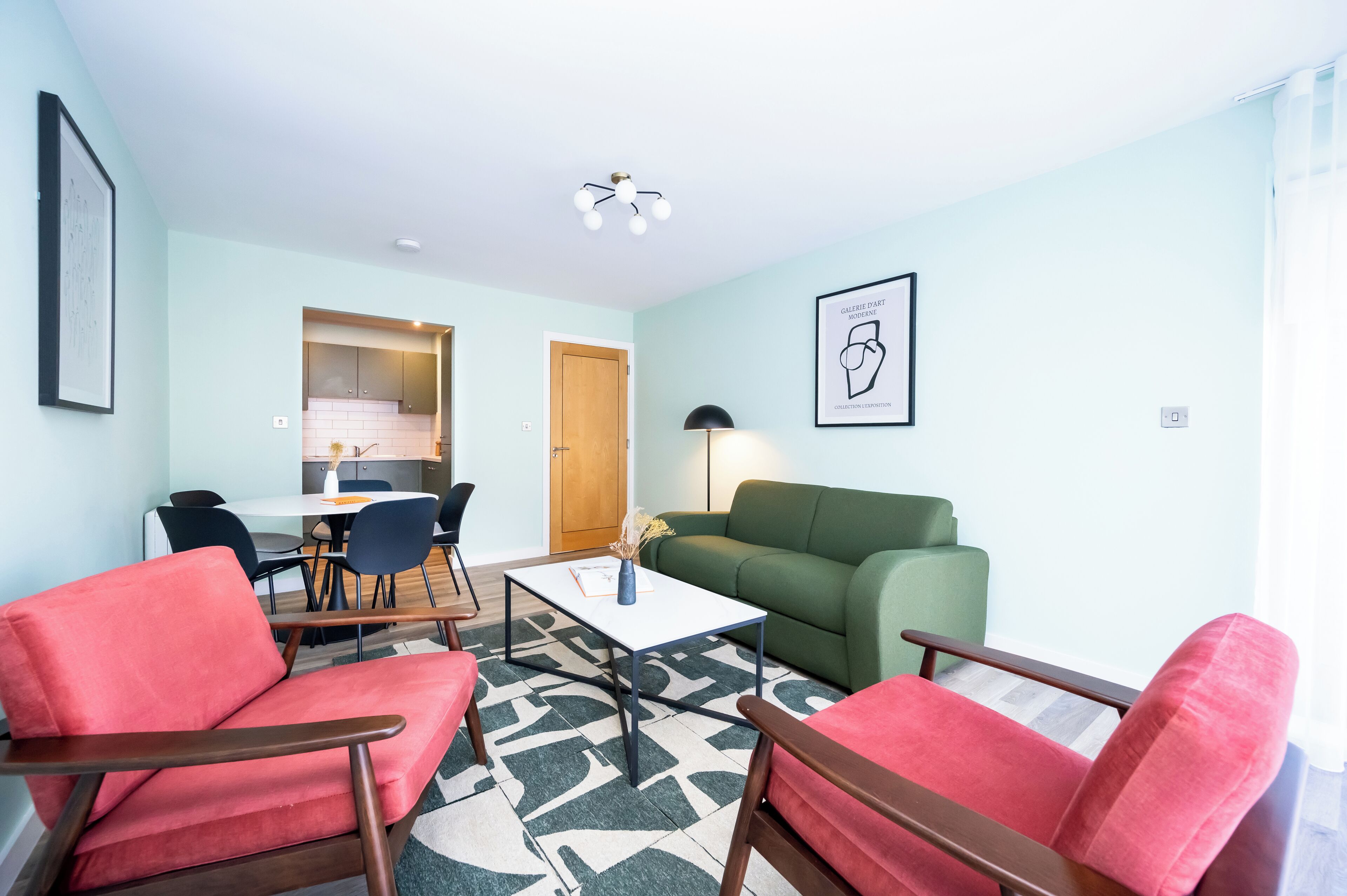 Una sala moderna con un sofá verde, dos sillones rojos, una mesa de centro blanca y un comedor con mesa y sillas.