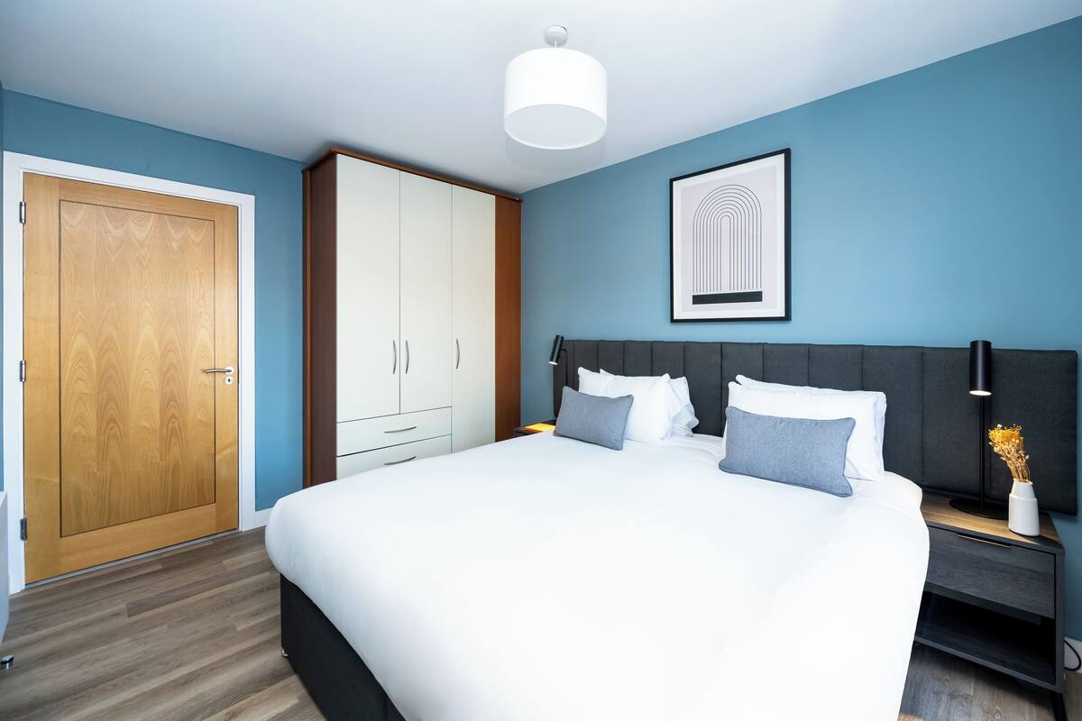 Een slaapkamer met een groot bed, een kledingkast, een nachtkastje en een ingelijste foto aan de wand.