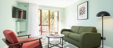 Een woonkamer met een groene bank, een rode fauteuil, een salontafel, een vloerlamp, een aan de muur bevestigde televisie en een groot raam met gordijnen.