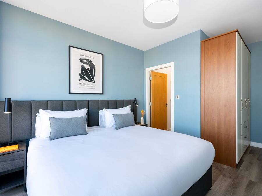 A neatly made bed with white and grey bedding, a framed artwork on the wall, a wooden wardrobe, and a nightstand with a lamp and a book.
