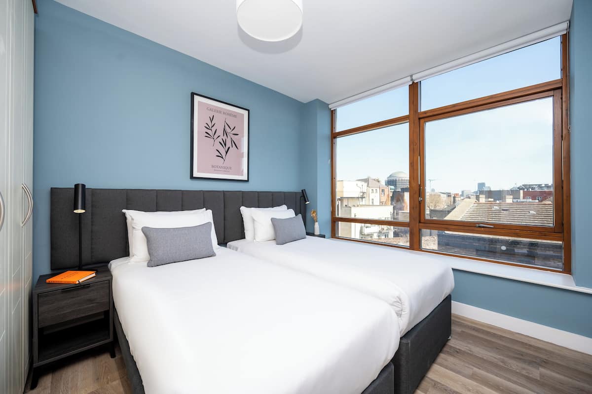 Een moderne slaapkamer met een groot bed, een nachtkastje, een botanisch kunstwerk in een lijst en uitzicht op de stad door een groot raam.