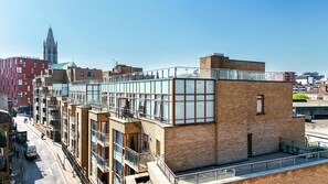 Un immeuble à plusieurs étages doté d’une terrasse sur le toit, avec en arrière-plan le clocher imposant d’une église, et un ciel dégagé.
