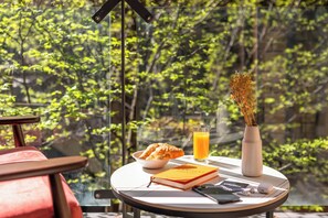 Une table de petit-déjeuner avec des croissants, un verre de jus d’orange, un livre, un vase de fleurs séchées et un téléphone intelligent, le tout sur un balcon donnant sur une forêt.