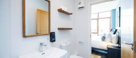 A modern bathroom with a white sink, toilet, and mirror. There are wooden shelves above the toilet and a window in the adjacent room.