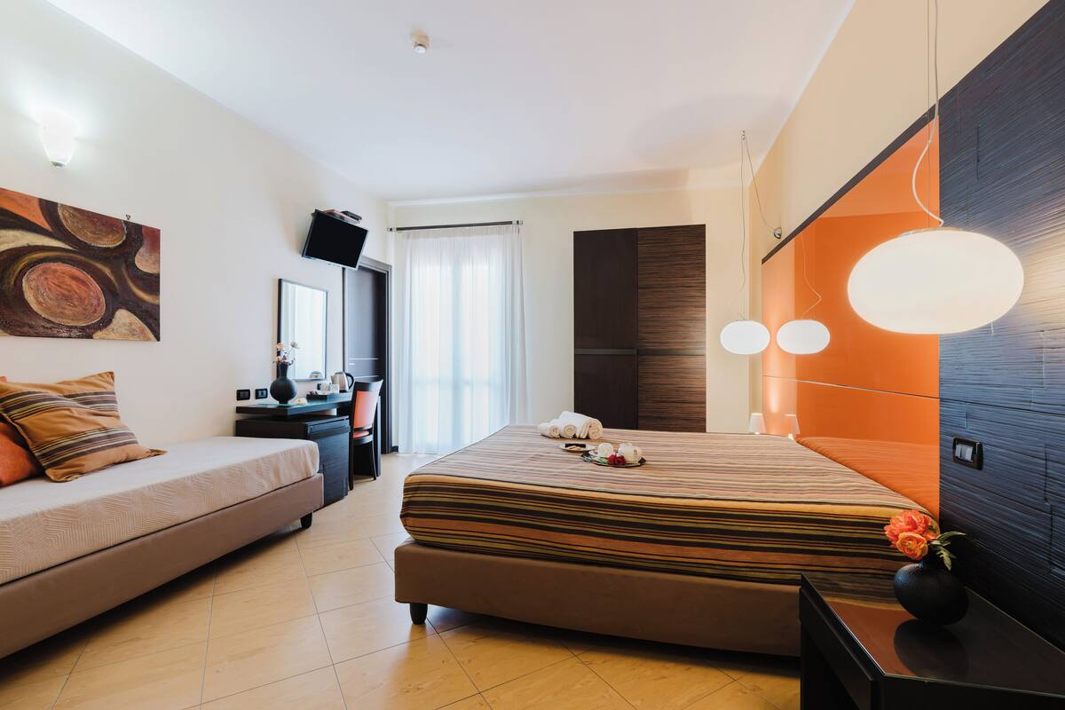 A modern hotel room with a bed, a sofa, a flat-screen TV, a desk, and a vase with flowers.