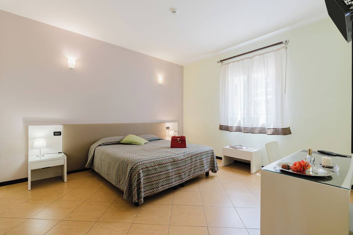 A bedroom with a bed, bedside table with lamp, desk with chair, and window with curtains.