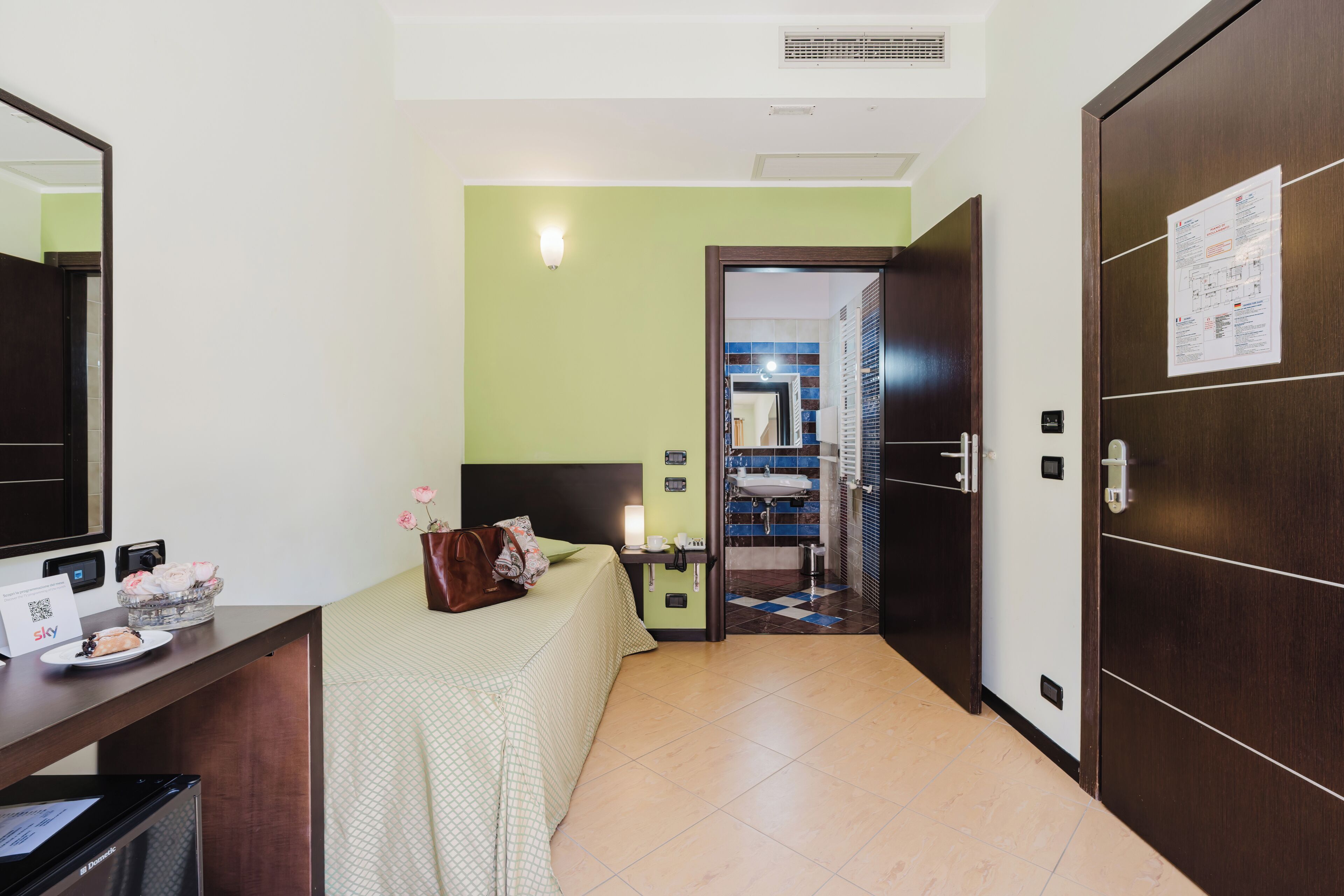 Une chambre d’hôtel avec un lit, une table de chevet et un bureau avec un ordinateur. On peut apercevoir la salle de bain à travers une porte ouverte.