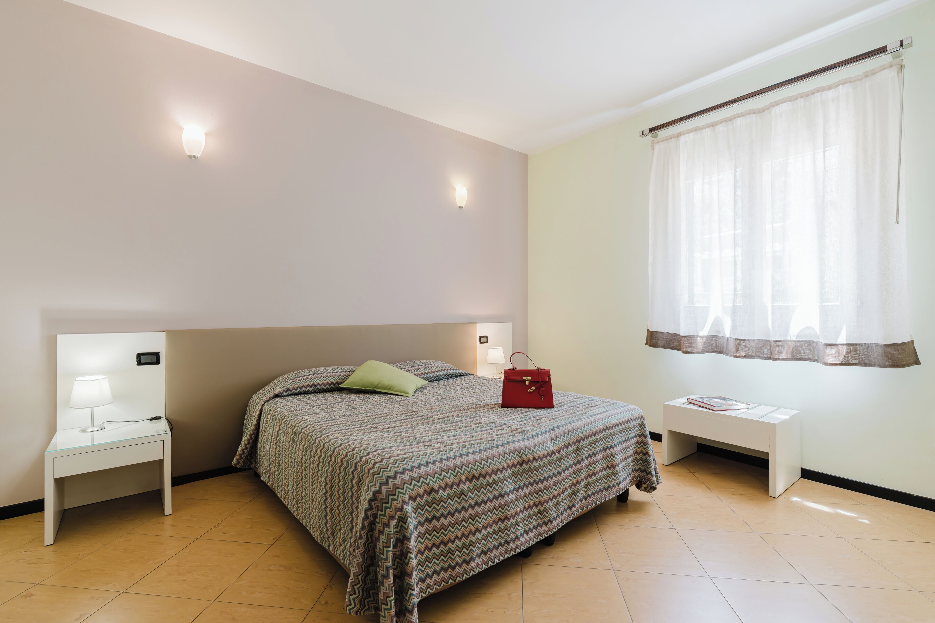 Quarto com cama, mesas de cabeceira, luminária na parede e cortinas na janela.