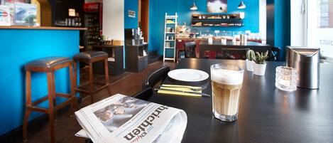 A bar area with wooden stools, a table with a newspaper, a glass of beer, and a metal container.