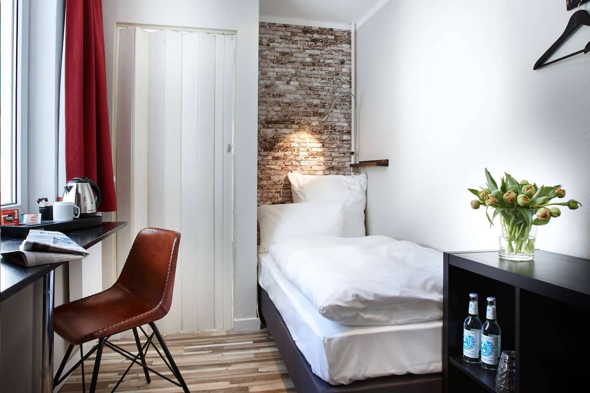 A hotel room with a brick accent wall, a bed with white linens, a red chair, a desk with a kettle, and a vase with flowers.